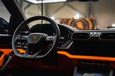 Lamborghini Urus 'un içini kapatın. Direksiyon simidini logo, İtalyan bayrağı aksanı, karbon fiber gösterge paneli ve turuncu detaylarla sergi salonunda gösteriyor..
