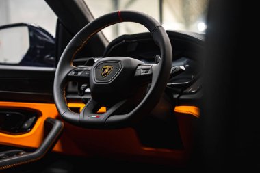 Lamborghini Urus 'un iç görünüşü, amblem, kırmızı şerit, İtalyan bayrağı detayı ve turuncu dikişli bir sergi salonunda direksiyona odaklanıyor..