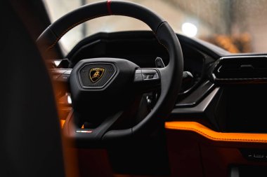 Lamborghini Urus 'un içinin yakın görüntüsü kırmızı dikişli direksiyonu, Lamborghini logosunu ve turuncu aksanlı ışıklandırmalı paneli gösteriyor..