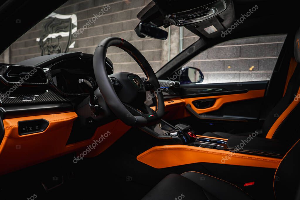 Close up of Lamborghini Urus interior featuring carbon fiber details, orange stitching, Italian flag emblem, and a concrete wall with Lamborghini logo.