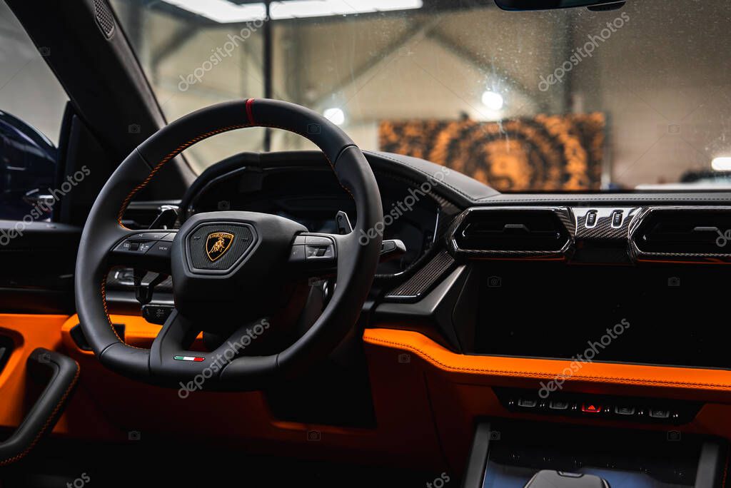 Close up of Lamborghini Urus interior showing the steering wheel with logo, Italian flag accent, carbon fiber dashboard, and orange details in a showroom.