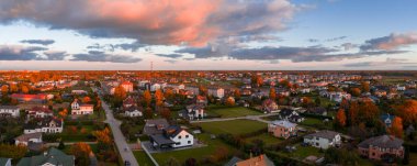 Panoramic aerial view of Marupe district in Riga, Latvia, with golden autumn foliage, neat houses, a red and white tower, and dramatic cloudy skies.