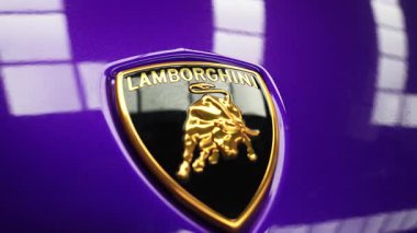 Lamborghini logosunun canlı mor bir yüzey üzerindeki detaylı görüntüsü altın boğa amblemi ve tepedeki ışığın parlak yansımalarını gösteriyor..