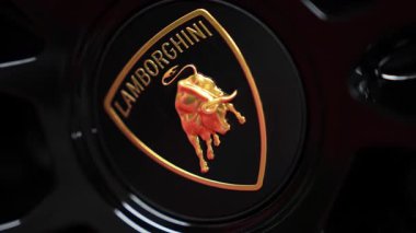Lamborghini logosunun altın boğa amblemi ve metnin yer aldığı parlak siyah bir yüzey üzerindeki detaylı görüntüsü. Kamera döner ya da hafifçe yakınlaşır.