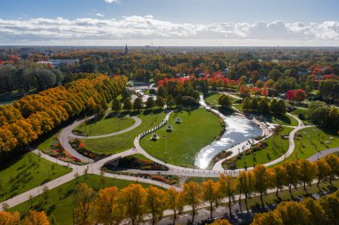 Riga, Letonya 'daki Zafer Parkı' nın havadan görünüşü, altın ve kırmızı sonbahar yaprakları, yansıtıcı bir gölet, kıvrımlı yollar ve bir şehir manzarası içeriyor..
