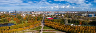 Riga, Letonya 'daki Zafer Parkı' nın havadan görünüşü, sonbaharda altın yapraklar, ağaç çizgili yollar, bir gölet, Daugava Nehri ve şehir simgeleriyle.