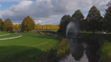 Riga parkının hava geçidinde yeşil çimenler, dolambaçlı yollar ve bir çeşme görünüyor. Bir dönme dolap ve sivri uçlu bir bina. Gün ortasında ufuk çizgisini işaretle..