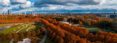 Hava görüntüsü sonbaharda portakal ağaçları, patikalar, çimenler ve bir spor sahasının olduğu bir Riga parkı gösteriyor. Ulusal Kütüphane ve şehrin yüksek binaları öğleden sonra Daugava 'nın ötesinde yer alır..