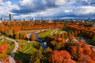 Öğleden sonra bir Riga parkının hava görüntüsü, dolambaçlı yollar, çimenler, bir su yolu, kırmızı ve turuncu ağaçlar, dönme dolap ve yüksek gökdelenli bir ufuk çizgisi..