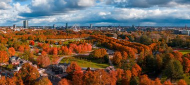 Riga, Letonya 'nın Ekim ayındaki hava görüntülerinde parklar, kırmızı ve altın ağaçlar, Işık Kalesi, Vansu Köprüsü, yüksek binalar, kilise kuleleri ve öğleden sonra dramatik bulutlar görülüyor..