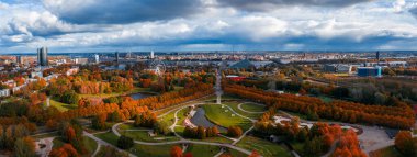 Aerial view of Riga, Latvia shows Kronvalda or Uzvaras Park with paths and ponds, Vansu Bridge, National Library, Old Town spires, and a riverside Ferris wheel in fall