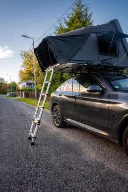 Sessiz bir banliyö caddesinde koyu gri lüks SUV parkları, alüminyum merdivenli çatı üstü çadırı. Sıcak alçak güneş ve geniş açı, yakınlardaki modern evleri ve boyutu vurguluyor..