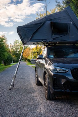 Ağaçlık bir yolda park edilmiş koyu renkli lüks bir cip, çatı çadırı açık ve merdiven genişletilmiş, öğleden sonra aydınlatması ön fasya ve kıyı boyunca çadır kapısı.