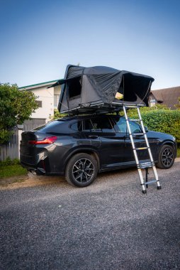 Çitlerin ve mütevazı evlerin yanına park edilmiş siyah bir BMW geçidi, teleskopik merdivenle açık bir çatı çadırı, öğleden sonra ışığı rahatlatıcı bir tatil havası yaratır.