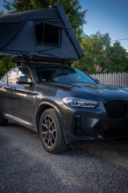 Koyu gri renkli lüks bir SUV, çakıllı bir garaj yolunda, erkek çizgileri ve yan camı açık bir çatı çadırında duruyor. Öğleden sonra ışığı ızgaraya ve çitin yanındaki tekerleklere yansıyor..