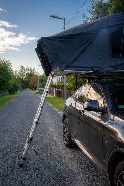 Çatı çadırı ve merdiveni olan parlak siyah bir SUV sakin bir iskan caddesinde oturuyor. Ilık gün ışığı evlere ve sokak lambalarına yansıyor..