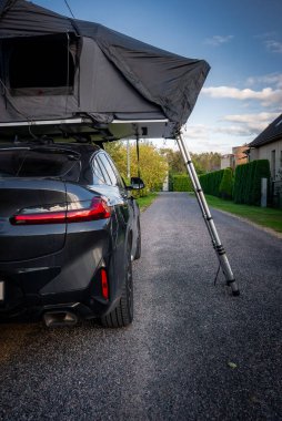 Banliyöde, çatıda çadır, çakıllı merdiven, arka taraf manzaralı, kırmızı LED arka far şeridi ve yumuşak öğleden sonra ışığında çift egzozlu siyah bir SUV park eder..