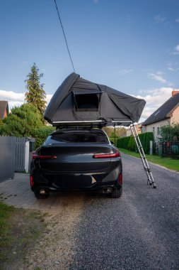 Siyah lüks bir SUV sakin bir banliyö caddesinde oturuyor, gri çatısı açık tente ve merdivenle asfalt döşeli, altın saat ışığı ve mavi gökyüzü rahatlamış seyahat havasını öneriyor..