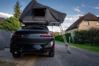 Banliyö caddesinde arka tarafında gri bir çatı çadırı ve merdiveni olan siyah lüks bir SUV park ediyor. Öğleden sonra ışığı, çitlerin ve çatıların yanında yumuşak bir kontrastı yansıtıyor..