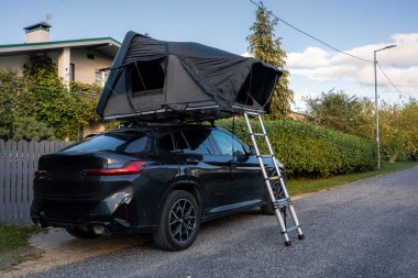 Sert kabuklu çatı çadırı ve merdiveni olan siyah lüks bir SUV, gece ışığında gri çitleri olan modern bir evin yakınındaki çitli banliyö sokağında park halinde..