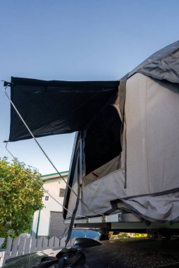 Close up shows a rooftop tent on a luxury SUV with open entry and awning, set near homes and a white picket fence, in warm daylight, suggesting relaxed travel.