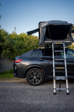 Koyu gri renkli lüks bir SUV, sahil kenarında çatı çadırı ve merdiveni olan bir park yeri. Öğleden sonra ışığı, temiz hatlara yumuşak gölgeler ekliyor ve kamp kurmaya hazır hale getiriyor..