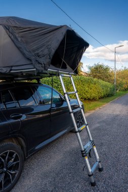 Çitlerin yanındaki sessiz bir yol kenarında siyah bir SUV park ediyor. Teleskop merdiveni olan bir çatı üstü çadırı açık. Öğleden sonra ışığı ve kapalı açılı görünüm form ve işlevi vurguluyor.