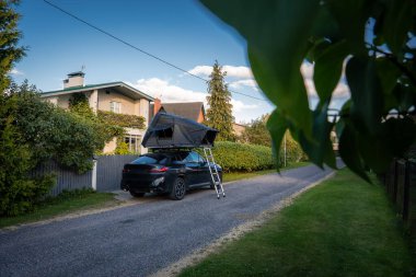 Sahil kenarındaki sessiz bir banliyö şeridinde uzanan çatısı ve merdiveni olan siyah lüks SUV, sıcak bir öğleden sonra ışığı, çitin arkasındaki çit ve çitli ev.