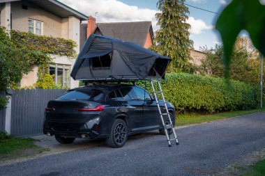 Sessiz bir banliyö sokağında, çatısı açık bir çadır ve merdiveni kaldırıma çıkan siyah lüks bir SUV park ediyor. Öğleden sonra ışığı ve yumuşak bulutlar, hazır bir kaçış havası olduğunu gösteriyor..