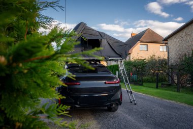 Çatı çadırı ve merdiveni olan siyah lüks bir SUV, öğleden sonra saatlerinde çitleri ve çitli bahçeleri olan sakin bir Avrupa konut şeridinde oturur.