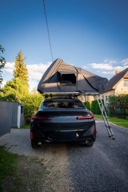 Banliyö caddesinde siyah bir SUV park ediyor, çatıdaki çadır merdiveni açık. Ilık gün ışığı çitlerin üzerinde taze gölgeler bırakıyor ve manzaralı çatı evleri sergiliyor..