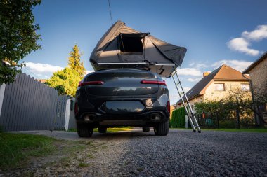 Koyu renkli lüks bir SUV, çatısı açık bir çadırı olan ve merdivenden inen bir patikaya park eder. Alçak güneş uzun gölgeler, düzenli evler ve bahçe çitleri sahneyi oluşturur..