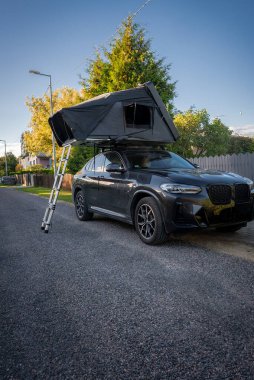 Sessiz bir banliyö sokağında çatısı ve merdiveni olan siyah lüks bir SUV park ediyor. Ahşap çitler ve ağaçlar çerçevelenmiş altın saat ışığı, taze bir portre.