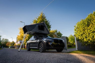 Altın saatinde sakin bir banliyö caddesinde siyah lüks bir SUV park eder, merdiveni açık çatı üstü çadırı, düzenli evler ve görülebilir uzun ağaçlar, geniş açılı ışık katar.