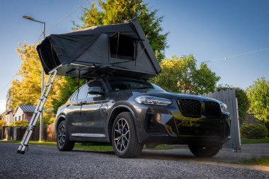 Çatı çadırı ve merdiveni olan koyu renkli lüks bir SUV sahil kenarındaki sakin bir yerleşim yerine park edilmiştir. Öğleden sonra ışıkları ve ağaçlar geniş açılı bir çerçeve oluşturuyor..