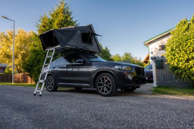Sessiz bir banliyö sokağında karanlık lüks bir SUV park ediyor, asfalta giden bir merdivenle siyah bir çatı çadırı açılıyor, ağaçların üzerinde sıcak bir gün ışığı ve yakınlardaki modern bir ev..