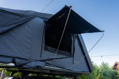 Close up of a rooftop tent on a luxury SUV near the ocean. Dark fabric, rainfly, mesh window, and taut lines are visible in bright daylight from a low angle.