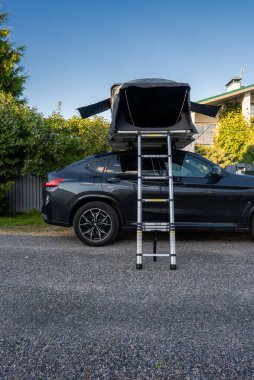 Çatı katı çadırı ve merdiveni olan siyah bir SUV bir çakıl taşının üzerinde oturur. Bir evin yakınına çekilir ve öğleden sonra ışığında bahçeler, tekerlekler ve sert kabuk tasarımı berrak görünür.