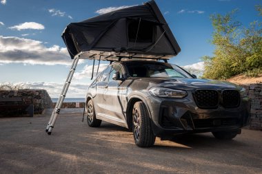 Koyu gri bir BMW X serisi SUV, alçak taştan bir duvarın yanına park eder, merdiveni açık bir çatı çadırı, ötesinde okyanus, sıcak altın saat ışığı ve dağınık bulutlar kamp yapmayı önerir..