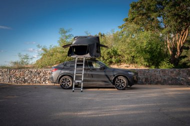 Çatı ve merdiveni olan koyu gri SUV çalıların ve büyük bir ağacın yanındaki taş duvarın yanında oturur. Gece yarısı ışığı uzun gölgeler oluşturur.