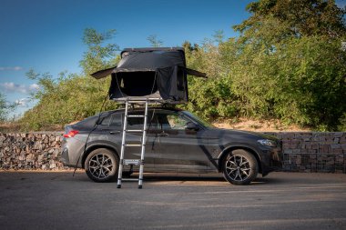 Kıyıdaki çalılıkların yanında alçak bir taş duvarın yanına park eden koyu renkli lüks bir SUV. Yan güneşlikleri ve merdiveni olan bir çatı çadırı. Öğleden sonra ışığı uzun gölgeler bırakır.