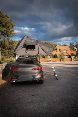 Ahşap renkli evlerin yanında gri lüks bir SUV parkı, alüminyum merdivenli çatı üstü çadırı. Evergreen ve kayalar arkada oturur, gün ışığı ağır bulutların arasından geçer..