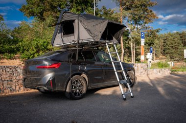 Çatısı olan koyu gri bir SUV, taş duvarın yanında çadır ve merdiven, kıyı şeridinde çam ağacı. Mavi park işaretleri ve sıcak gün ışığı bir yaz durağını işaret ediyor..