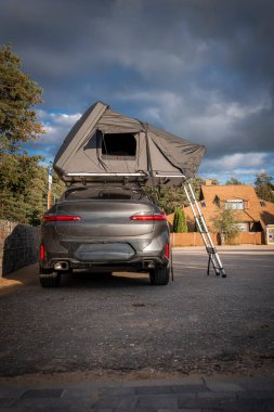 Gri lüks bir SUV ormanın yakınındaki banliyö kenarına park edilmiş, çatı üstü çadırı açık ve merdiven genişletilmiş. Çam ağaçları ve dağ evleri öğleden sonra aydınlığında ortaya çıkar..