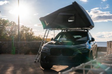 Koyu renkli bir SUV, taştan bir deniz kenarına park eder, çatı üstü çadırı açılır ve aşağı doğru merdiven iner, okyanusun üzerine alçak güneş ışığına bakar. Sıcak ışık ve uzun gölgeler öğleden sonra rahatlığını gösteriyor..