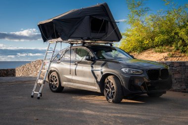 Kıyı manzaralı koyu gri SUV parkları, çatıdaki çadır merdiveni açık. Taş duvarlar, kumlu ovalar ve okyanus, gün ışığı ve engebeli kıyıları gösteriyor.