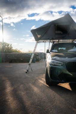 Koyu renkli lüks bir SUV, alçak taştan bir deniz kenarına park ediyor, çatı üstü çadırı açık ve merdivenler kaldırımda. Sıcak öğleden sonra ışığı okyanus ufkunun yakınında uzun gölgeler bırakır..