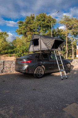 Düşük Gabion duvarlı, çatısı ve merdiveni olan koyu gri bir SUV park ediyor. Öğleden sonra ışığı metali ve kumaşı ısıtır. Uzun ağaçlar, kumlu toprak ve P işareti kıyı olduğunu gösteriyor..