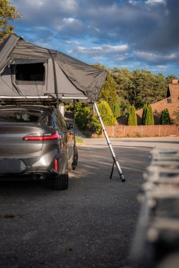 Tuğla binalar ve yüksek çitlerin yanındaki asfalt bir yolda park etmiş gri lüks SUV, öğleden sonra ışığı, alçak arka üç çeyrek açı, karamsar bulutlar..