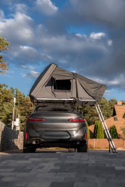 Gri lüks bir SUV asfaltlı bir garaj yolunda oturur, çatı üstü çadırı açılır ve merdiven iner, yakınlardaki tuğla evler ve kozalaklı ağaçlar, öğleden sonra ışığı ve kümülüs bulutları macera havası yaratır..